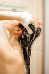 Beautiful naked young woman using shampoo while taking shower in bathroom
