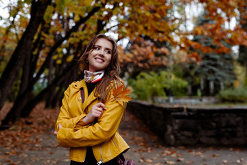 autumn portrait of a beautiful smile girl with long fair-haired hair in yellow leather jacket and a maple leaf in hands