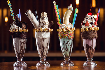 Four unique giant milk shakes in row on a wooden table.