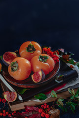 Dark autumn still life, ripe persimmons on a wooden plate with autumn leaves. Seasonal fruits close-up. Dark food photography with copy space.