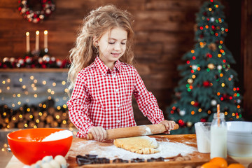 Happy little child, cute kid girl at the table in domestic kitchen making gingerbread xmas cookies decorated for Christmas holiday. Girl helping and having fun