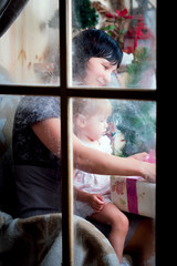 Cheerful mom and her cute daughter girl exchanging gifts. Parent and little child having fun near Christmas tree indoors. Loving family with presents in room.