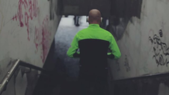 Steadicam back shot of adult bold sportive man jogging in the city on a rainy autumn day. Regular workout with favorite music. Jogger working out in the morning outdoors, rear view.