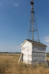 A vintage wooden pump house with white peeling paint and remnants of a wind mill on top, photographed from the side in natural light.