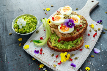 Tasty sandwich with avocado boiled eggs, pumpkin seed and edible viola flowers in a white board. healthy food