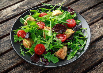 Fresh salad with chicken breast, arugula, nuts and tomatoes on black plate in a wooden table.
