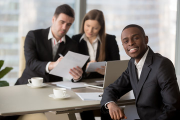 Portrait of confident african american millennial businessman sitting at desk, caucasian colleagues working on document at background. Multinational financial analysts examining company annual report