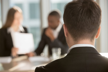 White male job candidate waiting for employers decision, multinational HR managers analyzing resume on background. Man in formal wear interested in vacancy visiting job interview. Close up back view