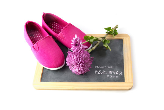 pink kid shoes and  flower on a writing blackboard isolated on white, german text Internationaler Maedchentag, that means International Day of the Girl Child 11 October, selected focus