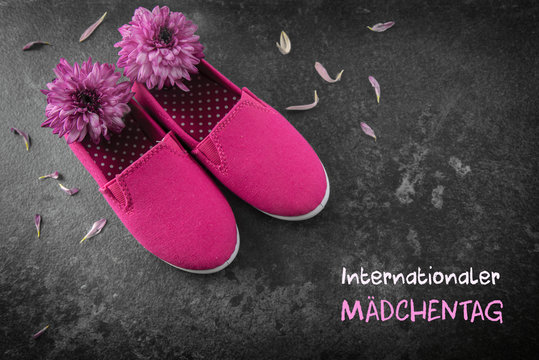 pink kid shoes and flowers on a dark slate background, german text  Internationaler Maedchentag, that means International Day of the Girl Child,  date concept 11 October, copy space