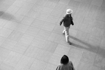 Man in a white hat and jacket and an Asian woman walk through a train station in Europe.  Looking down on two people from above, black and white image.