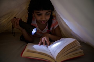 Girl holding flashlight while reading novel under blanket