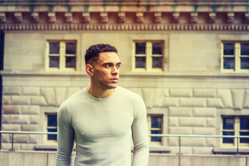Fototapeta premium Portrait of young handsome American Man in New York, wearing light gray, collarless knit pullover sweater, standing in front of vintage office building, looking away, thinking. Retro filtered effect.