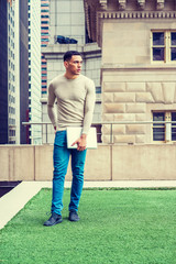 Way to Success. American Graduate Student studying in New York, wearing collarless knit sweater, jeans, sneakers, holding laptop computer, stands on green lawn on campus. High buildings on background.