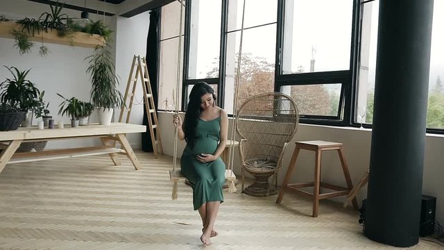 Young pregnant girl swinging on a wooden swing. A young girl in a long green dress on a swing in the studio.