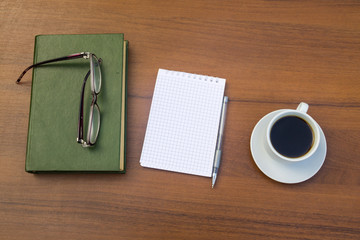 Cup of coffee, book, notepad, pen and eyeglasses on wooden desk