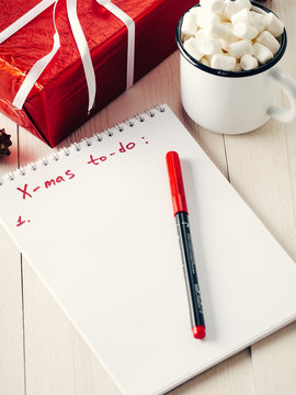 Christmas to-do list. Xmas gifts shopping planning. Make shopping or to-do list for Christmas. Notebook, mug hot chocolate with marshmallows and New Year's gift on white wooden background.