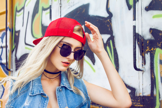 Beautiful young blonde girl dressed in a street style outfits posing in front of wagon full of graffitis. Wearing red cap, black sunglasses and choker around her neck with triangle.