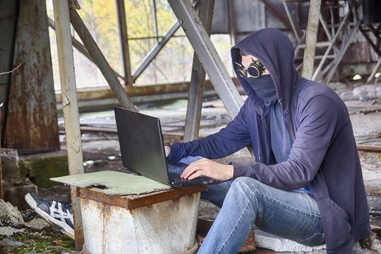 Cyberpunk hacker or programmer or cyber terrorist in goggles with spikes, mask and hood working on a laptop among the apocalyptic landscape of old rusty half-destructed building. HDR