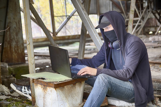 Cyberpunk hacker or programmer or cyber terrorist in cap, mask and hood working on a laptop among the apocalyptic landscape of old rusty half-destructed building. HDR