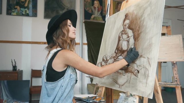 Caucasian female artist, in a black smart hat, drawing the sketch of a woman in her studio, slow motion