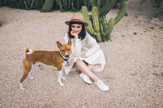 Happy brunette girl walks in cacti park in old fashioned white dress and brown hat with her clever basenji dog
