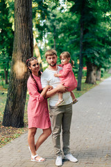 Fototapeta premium young happy family with little beautiful baby with blue eyes walking in summer park at sunset. Mom and little daughter in identical pink dresses