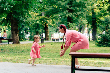 Obraz premium young happy family with little beautiful baby with blue eyes walking in summer park at sunset. Mom and little daughter in identical pink dresses