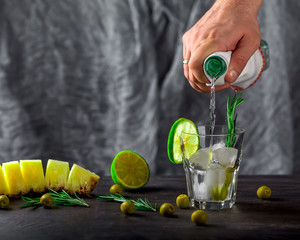Hand pours alcohol into a glass with ice. Olives, lime and rosemary in a glass and nearby