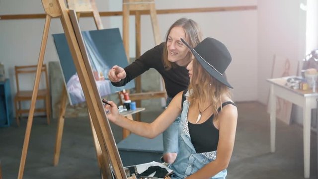 Male artist, in casual torn jeans, consulting his female colleague, in balck hat and denim dangarees, while drawing, slow indoor motion