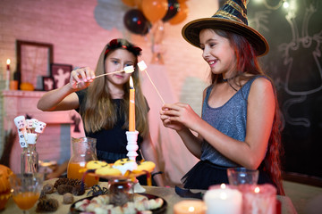 Two joyful friends wearing Halloween costumes making fried marshmallows with help of candle flame while enjoying home party, interior of decorated room on background