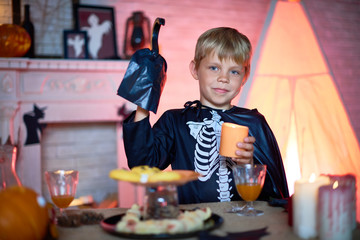 Cute little boy wearing skeleton costume holding candle in hand while looking at camera with warm smile, interior of living room decorated for Halloween party on background