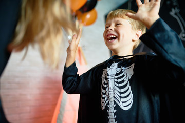 Waist-up portrait of laughing little boy wearing skeleton costume enjoying Halloween home party,...
