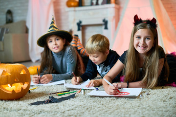 Group portrait of cheerful children wearing Halloween costumes gathered together at cozy living room decorated for Halloween and drawing colorful pictures, girls looking at camera with wide smiles