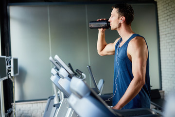 Concentrated sportsman in headphones refreshing himself with water while running on treadmill at...