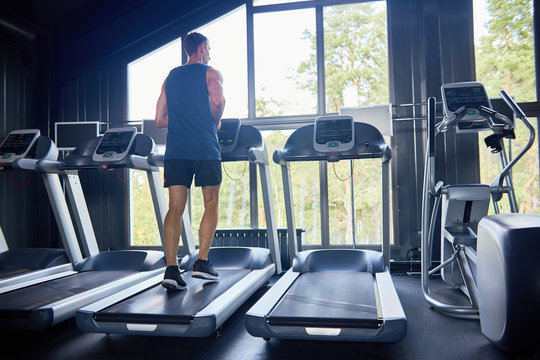 Back view of muscular athlete doing exercise on treadmill and enjoying picturesque view through panoramic windows of modern gym