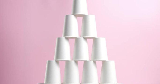 Young woman builds and breaks tower pyramid of white paper cups turned upside down on pink background