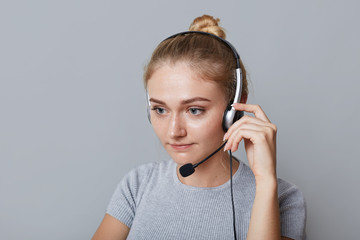 Serious female helpline operator uses headphones for her work, being focused on something, isolated over grey background. Businesswoman phones partners. Business, call centre and technology concept