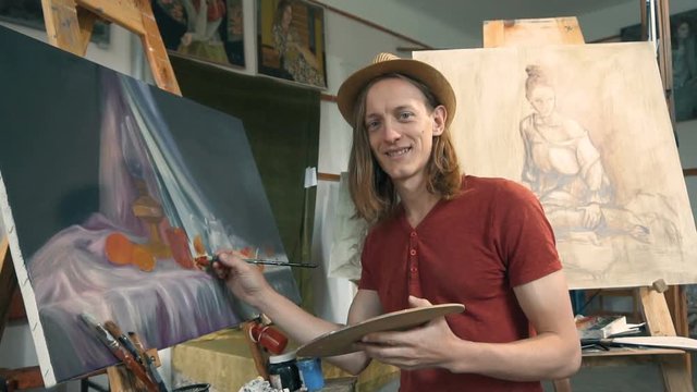 Long hair caucasian male artist, in a light brown hat, smiling during painting in the studio, slow motion