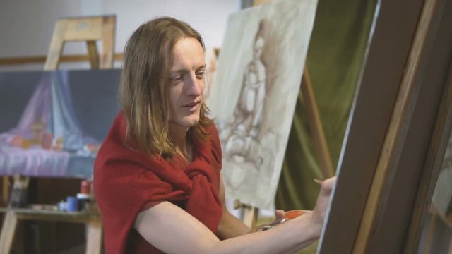 Long hair caucasian male artist smiles while painting a picture in the studio, slow indoor motion