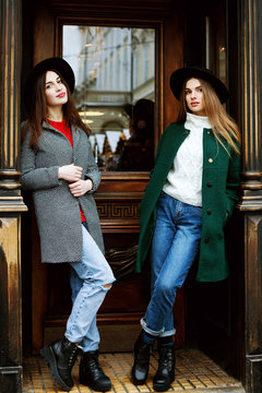 Outdoor Full Body Portrait Of Two Young Beautiful Fashionable Girls Posing Near The Door. Models Wearing Stylish Winter Warm Coats, Knitted Sweaters, Jeans, Boots And Hats. Female Fashion Concept