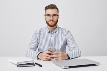 Horizontal portrait of unshaven pleasant looking young businessman sits at working desk, holds mobile phone as waits for important call, looks confidently into camera, isolated over white background