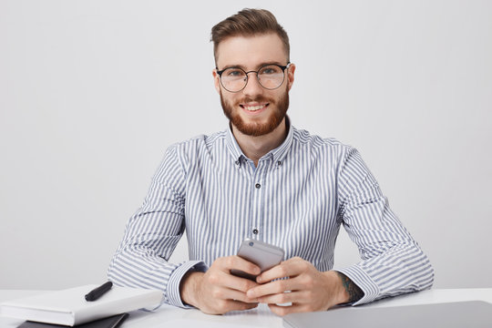 Headshot of good looking successful male manager waits for partners in office, holds smart phone, entertains as has spare time, surfe social networks or browse net. Modern technology concept