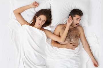 Indoor portrait of attractive Caucasian woman sleeping tight in bed lying on white sheets side by side with her bearded husband after hard working day at office. Tired couple seeing sweet dreams