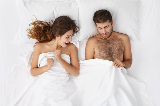 Picture of adult European bearded man and excited woman lying in bed and peeping under white blanket. Couple going to have sex in white bedrrom or hotel room. Love and relationships problems