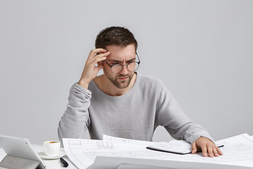 Clever male creative worker tries to concentrate on drawings, doesn`t understand where is his mistake, looks puzzled. Indoor shot of unshaven man in round spectacles being busy with making sketches