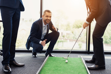 A respectable elderly man is playing golf in his office. Next to him are his workers. They look like the boss is playing