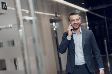 A respectable man in a strict suit walks along the office corridor with a phone in his hands, smiles and talks