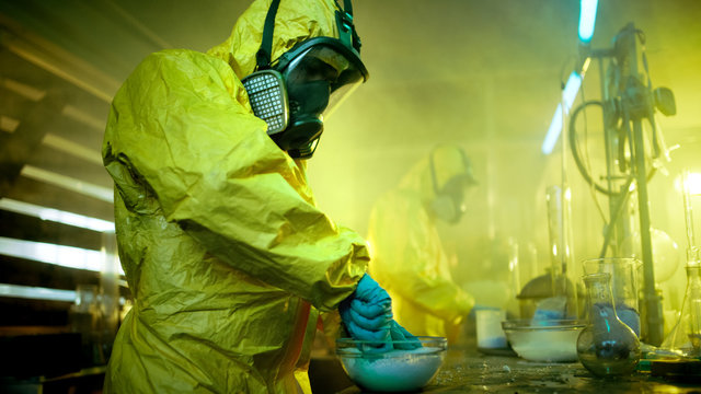 In the Underground Drug Laboratory Two Clandestine Chemists Cook Synthetic Drugs, Crush Them in a Bowl and Package for Further Distribution. They Wear Protective Gas Masks and Coveralls and Squat.