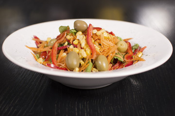 A warm salad with corn, pepper, olives, and lettuce on a white ceramic plate on a dark background. Selective focus. Tasty restaurant meal.